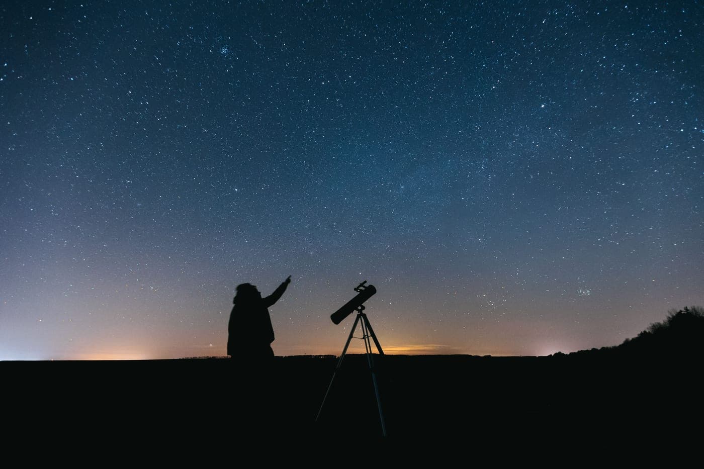A person silhouetted beside a telescope under a starry sky