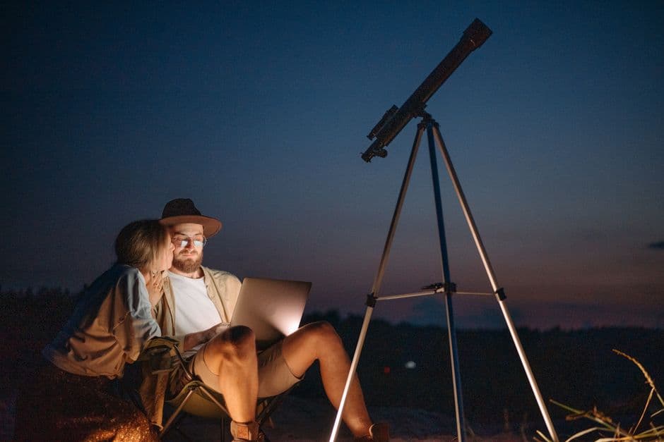 Two people stargazing with a telescope and a laptop at dusk
