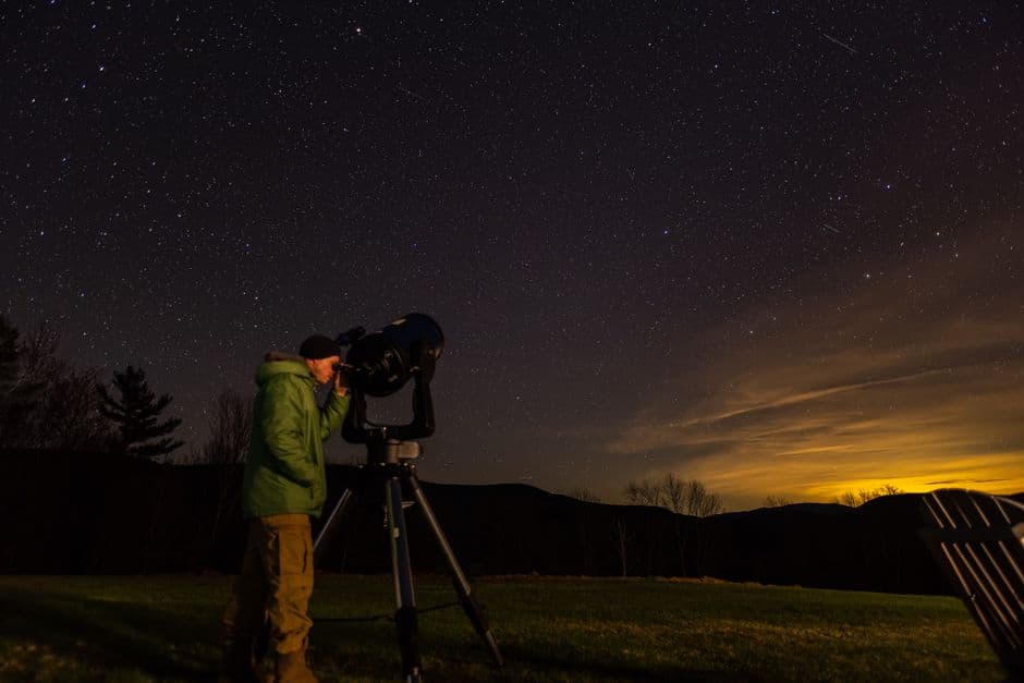 A person in warm clothing observing the night sky through a telescope on a grassy hillside