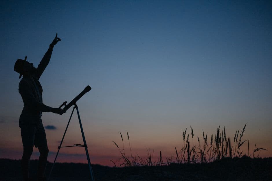 A person pointing at the sky beside a telescope at twilight