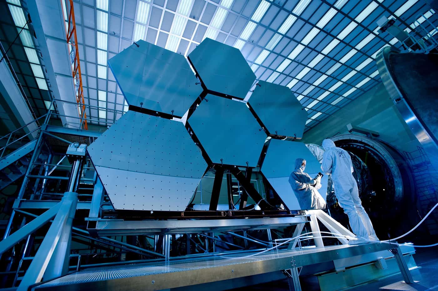 Engineers in protective suits working on a large telescope mirror in a laboratory