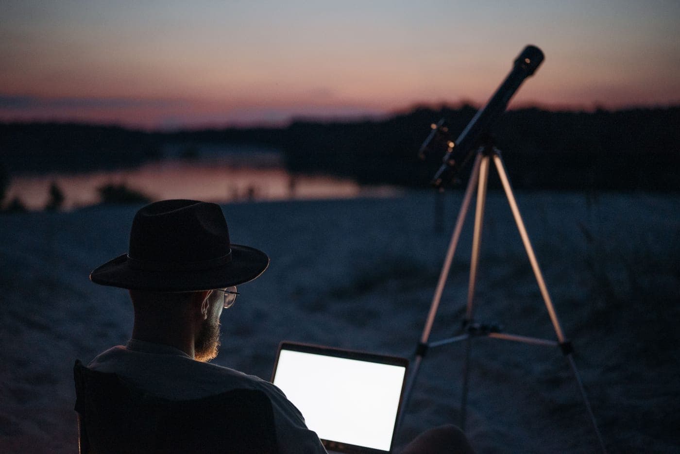 A person stargazing beside a lake at twilight with a telescope
