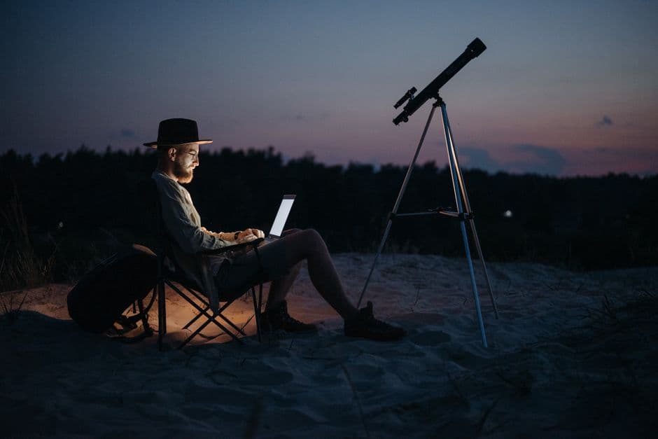 A camper at night beside a telescope and a laptop under a twilight sky
