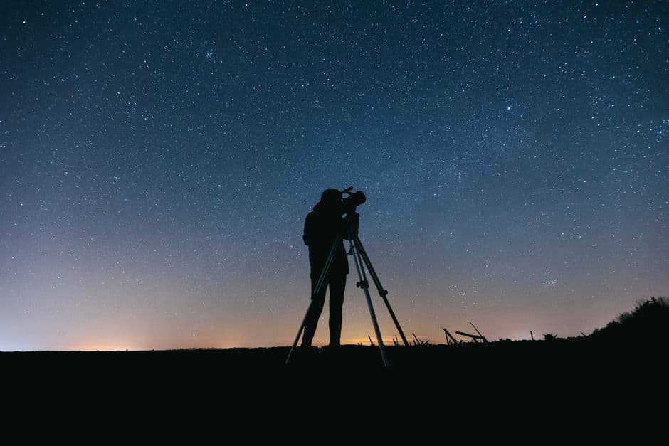 A person silhouetted at a telescope under a starry night sky